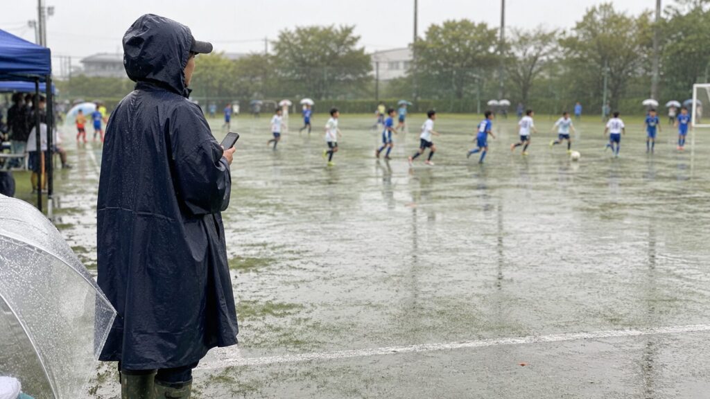 レインコートと長靴を着用し、雨の中で行われる子供のサッカー試合を見守る保護者