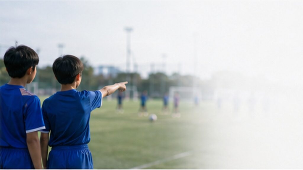 サッカーコートを背にした二人の少年が、片方の指す方向を見ながら立ち位置を確認する場面